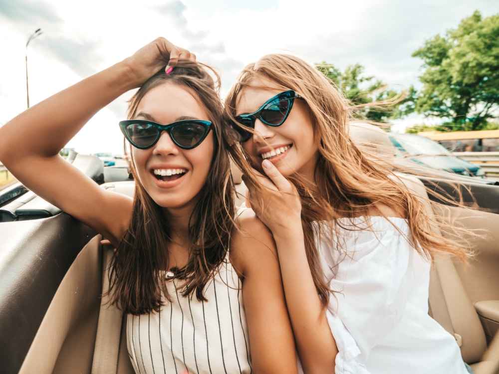 Portrait of two young beautiful and smiling hipster female in convertible car. Sexy carefree women driving cabriolet. Positive models riding and having fun in sunglasses